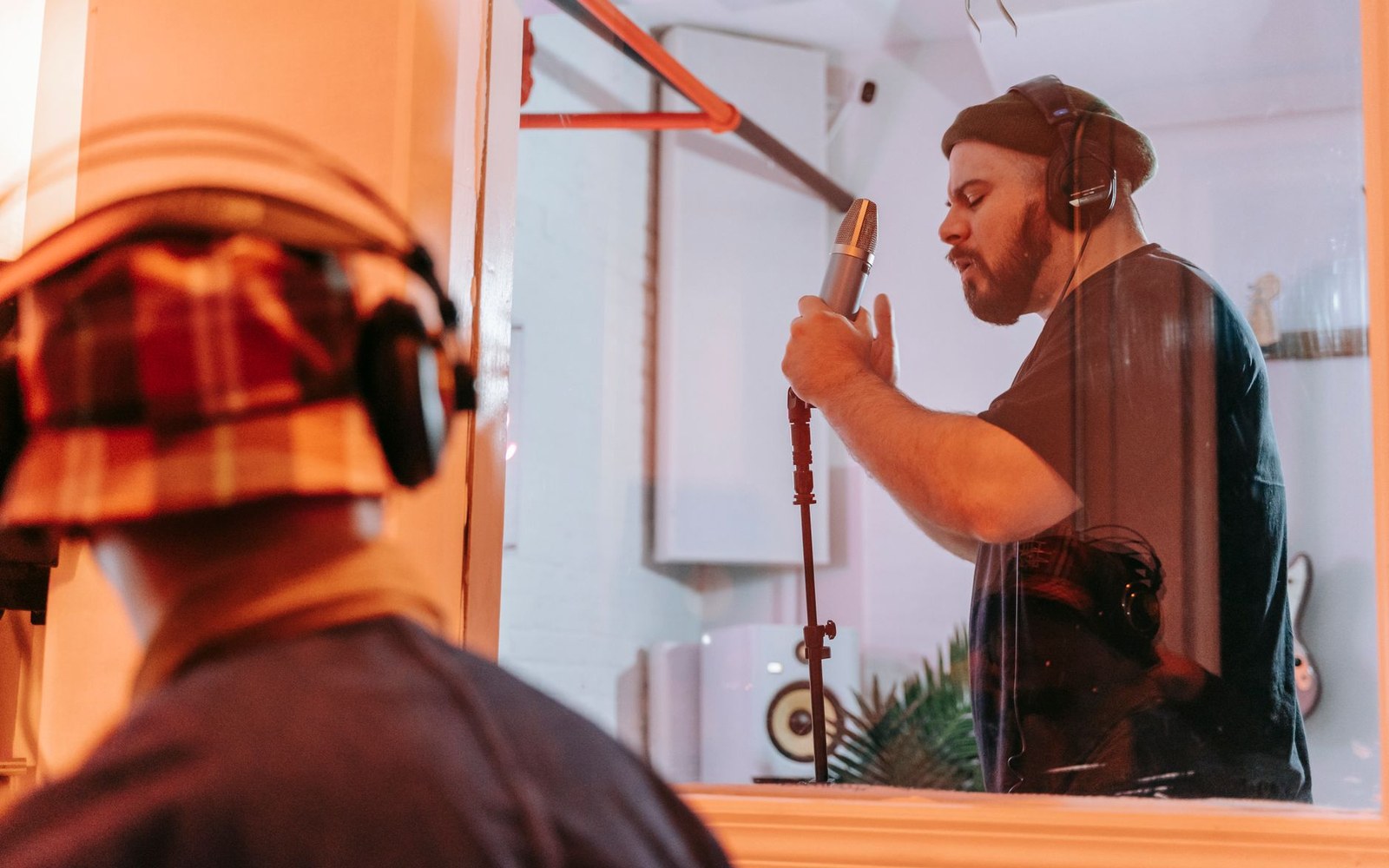 Adult vocalist at a studio microphone in a vocal booth, stage-lit, face visible through the glass